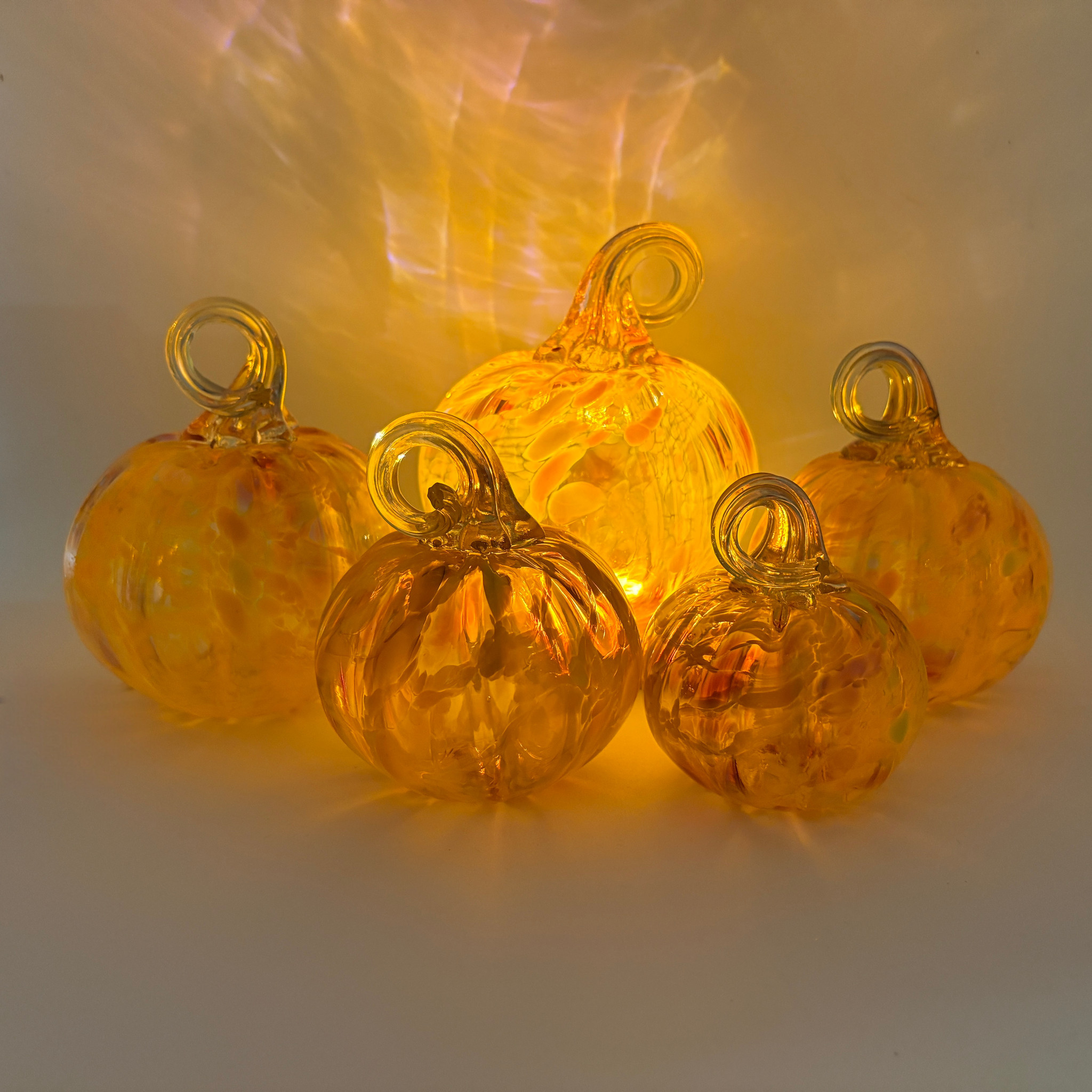 Five small transparent glass pumpkins with loops at the top, arranged together on a white surface. A warm yellow light shines through them, casting a glow on the background.