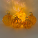 Five small transparent glass pumpkins with loops at the top, arranged together on a white surface. A warm yellow light shines through them, casting a glow on the background.