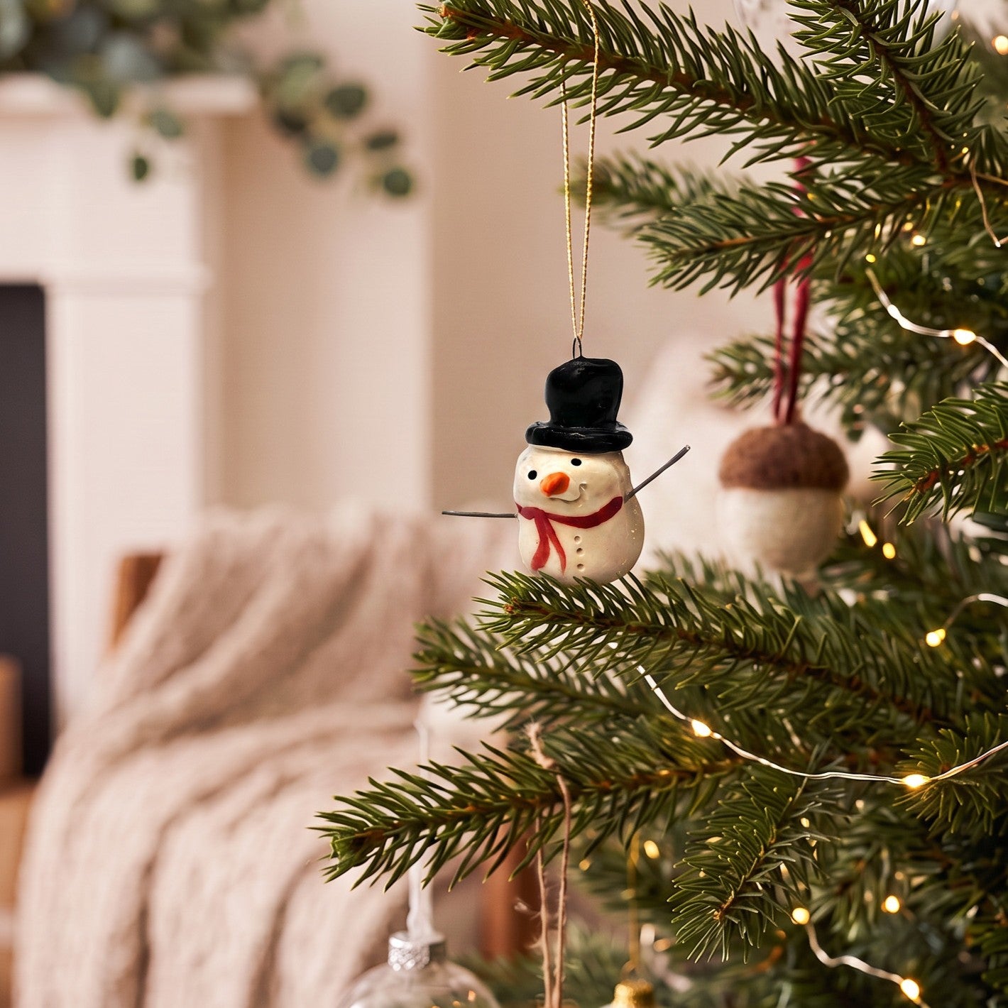 A small ceramic snowman Christmas ornament with a black top hat, red scarf, and orange carrot nose hangs on a green Christmas tree branch with warm string lights in a cozy indoor setting.