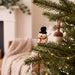 A small ceramic snowman Christmas ornament with a black top hat, red scarf, and orange carrot nose hangs on a green Christmas tree branch with warm string lights in a cozy indoor setting.