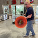 Glassblower Lisa Oakley in a workshop holding a large, bright red-orange hot disc of molten glass. The scene is the Oakley Hot Glass Studio with concrete floors, windows, and workshop equipment in the background.