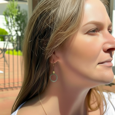 A close-up side profile of a woman wearing a delicate hoop earring with a textured lower half, set against an outdoor patio background.