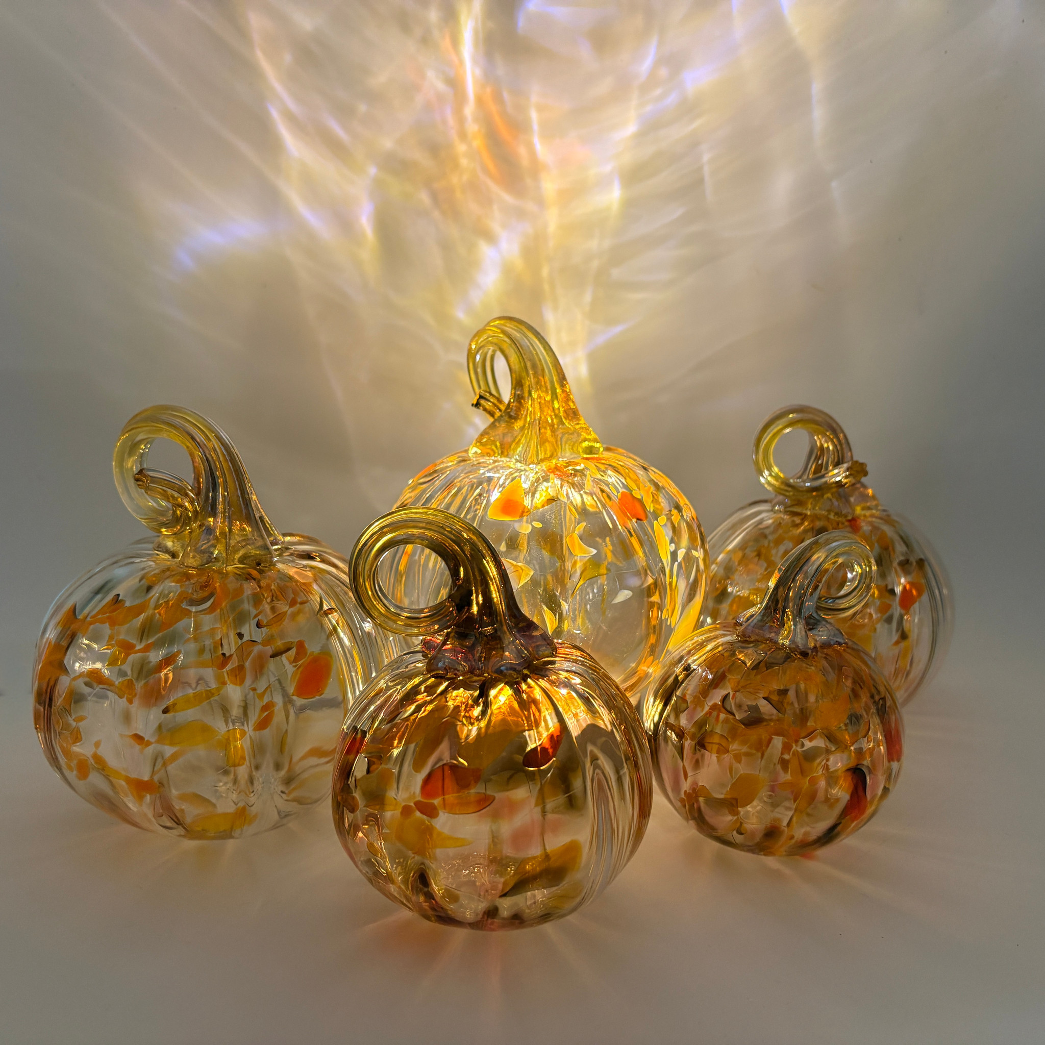 Five decorative glass pumpkins with ribbed textures and looped handles, filled with orange, yellow, and white marbled swirls, arranged on a reflective surface with dramatic light projections behind them.