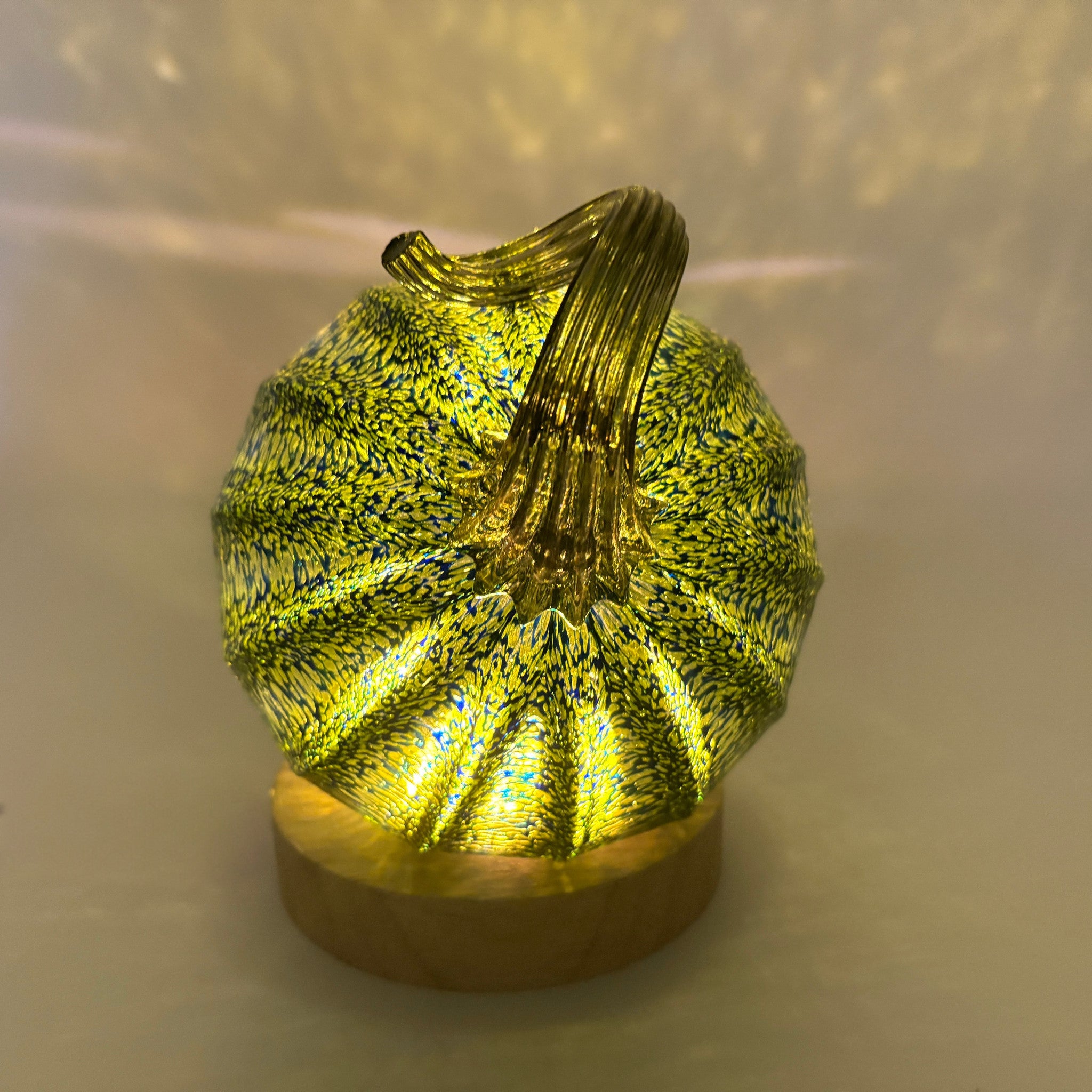 A handcrafted glass pumpkin with a ribbed body, a curved stem, and a warm yellow glow from within, sitting on an LED display light with a round wooden base. 