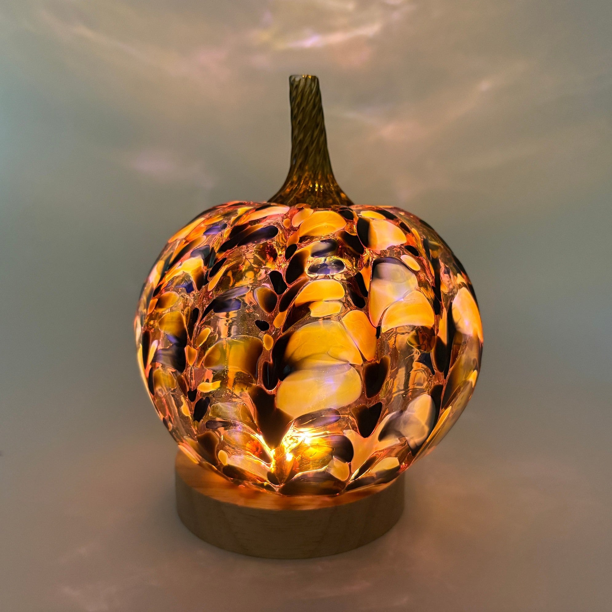 A handcrafted glass pumpkin with an illuminated marbled pattern in amber, gold, orange, and dark brown tones, sitting on an LED display light with a round wooden base. 