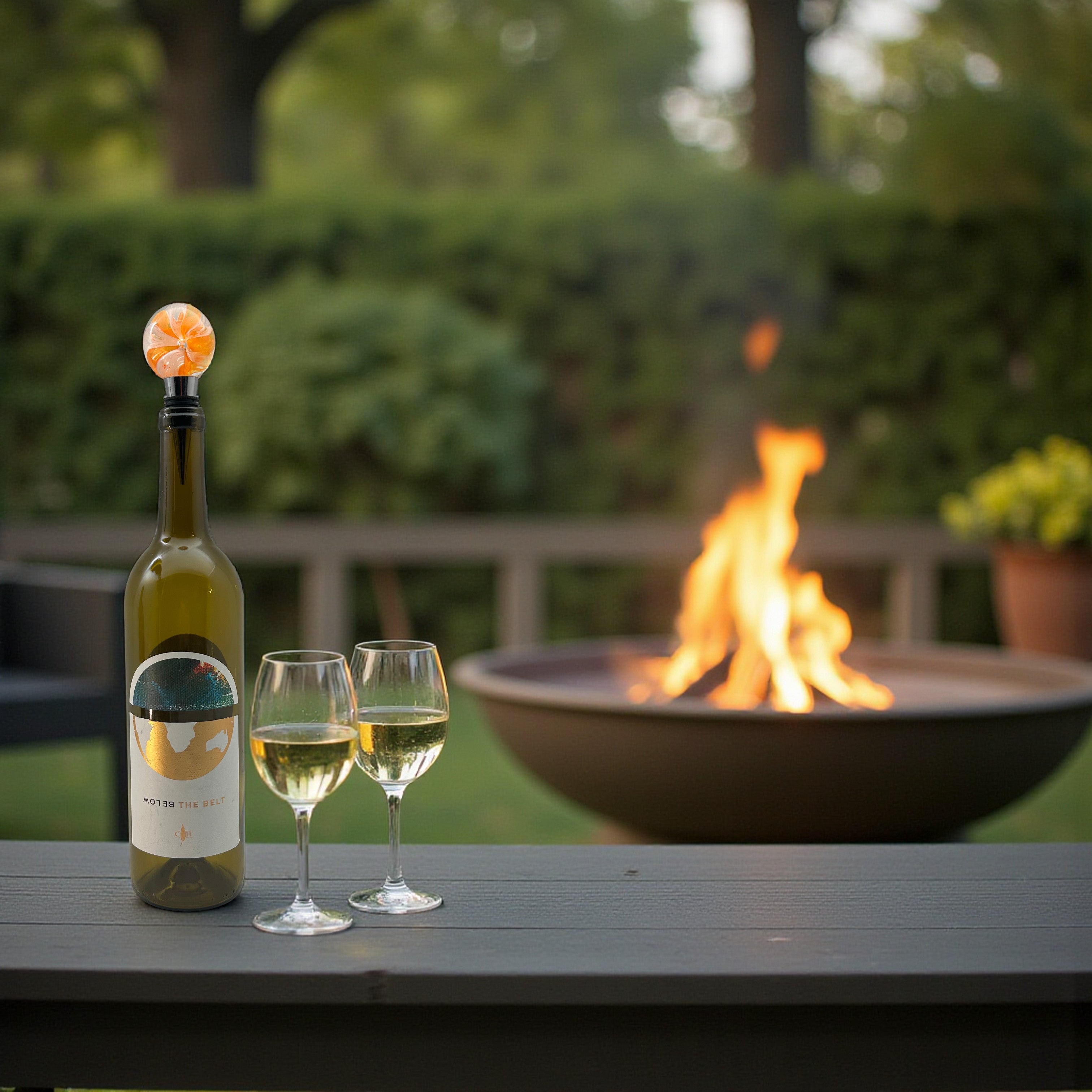 Wine bottle with a wine glass and another glass on a table with a fire pit in the background