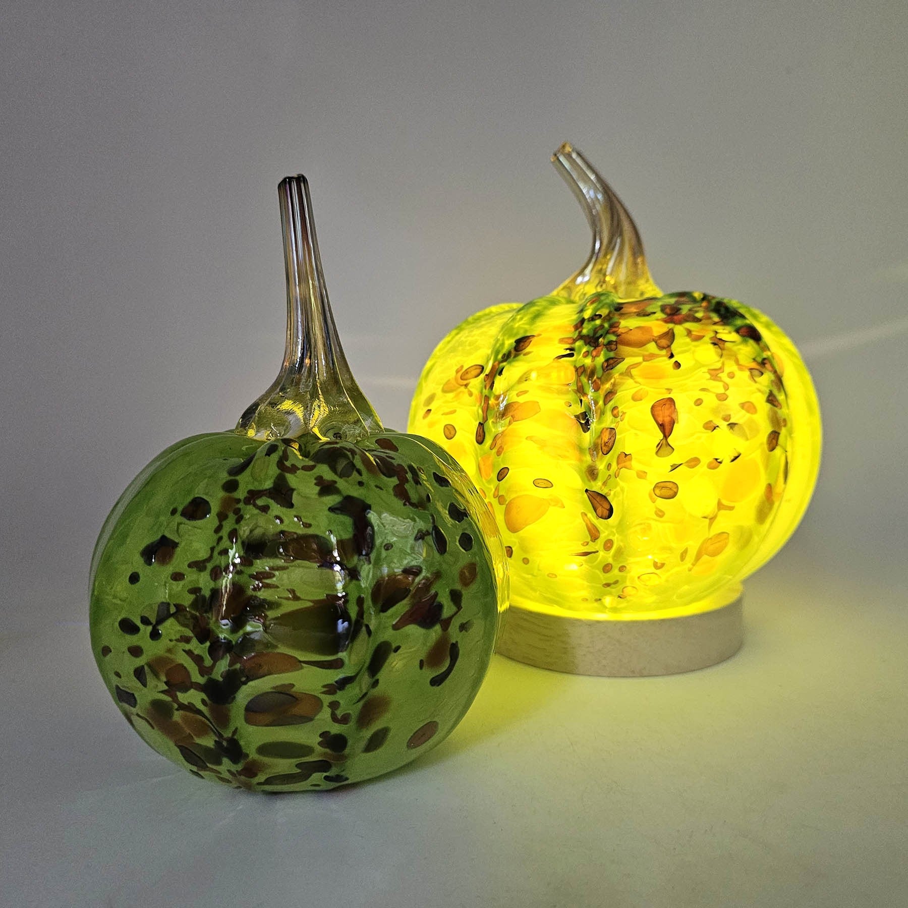 Two orchard glass pumpkins, one green and one yellow with brown speckles, on a gray background.