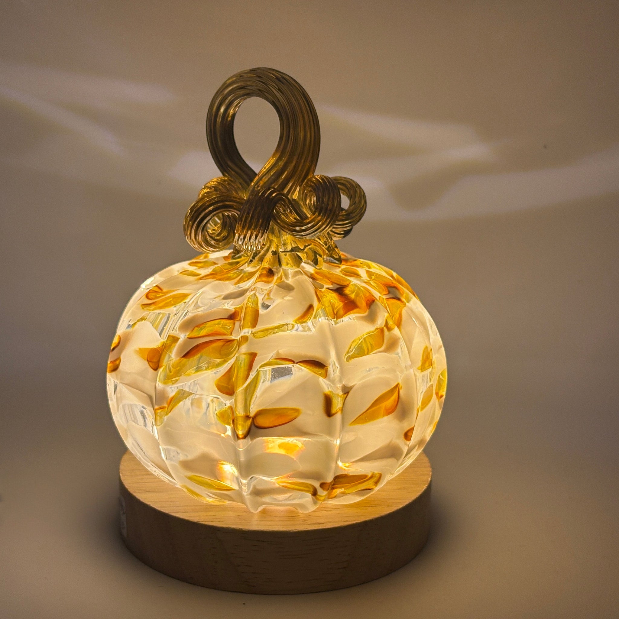 A handcrafted glass pumpkin with a warm white glow, featuring embedded yellow and orange leaf patterns, a dark brown glass stem loop, sitting on an LED display light with a round wooden base.