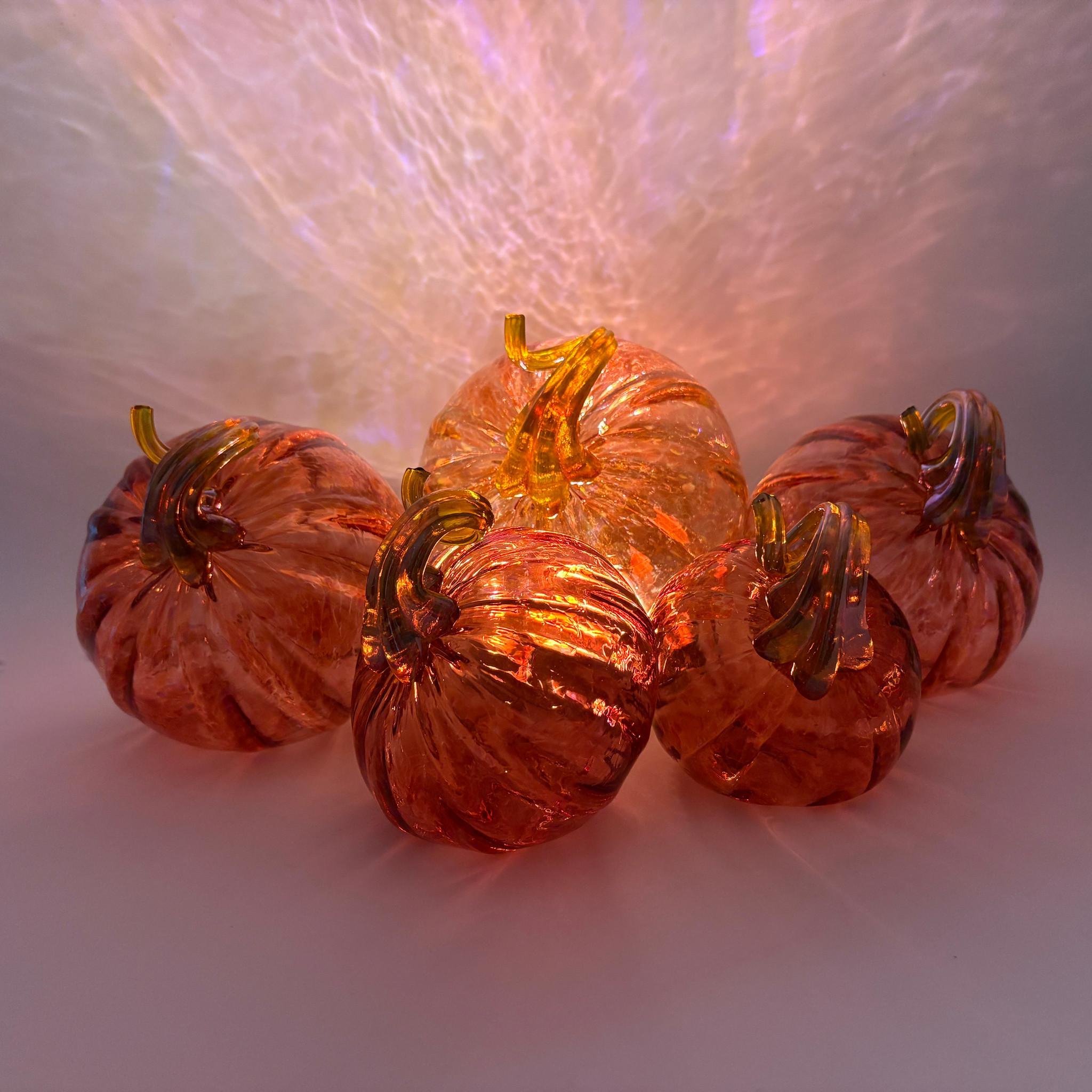 Five decorative glass pumpkins arranged together on a white surface, glowing with warm orange and amber light from within. The pumpkins have ribbed textures and translucent amber glass with metallic gold-toned stems.