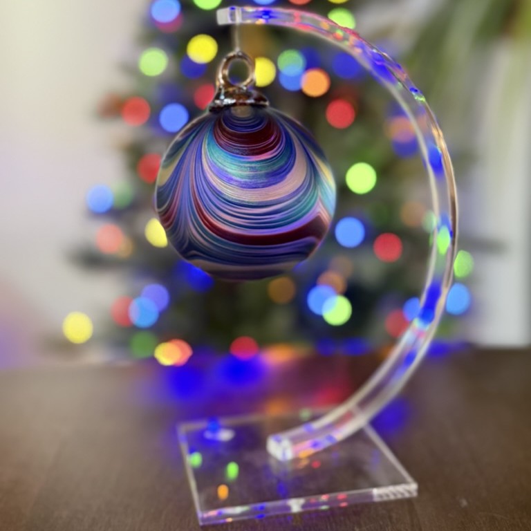 Colorful marbled Christmas ornament on a clear stand with blurred Christmas tree lights in the background.