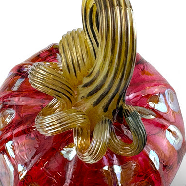 A close-up of a hand-blown glass pumpkin with a textured red body and a ribbed amber-yellow stem featuring dark brown stripes.  