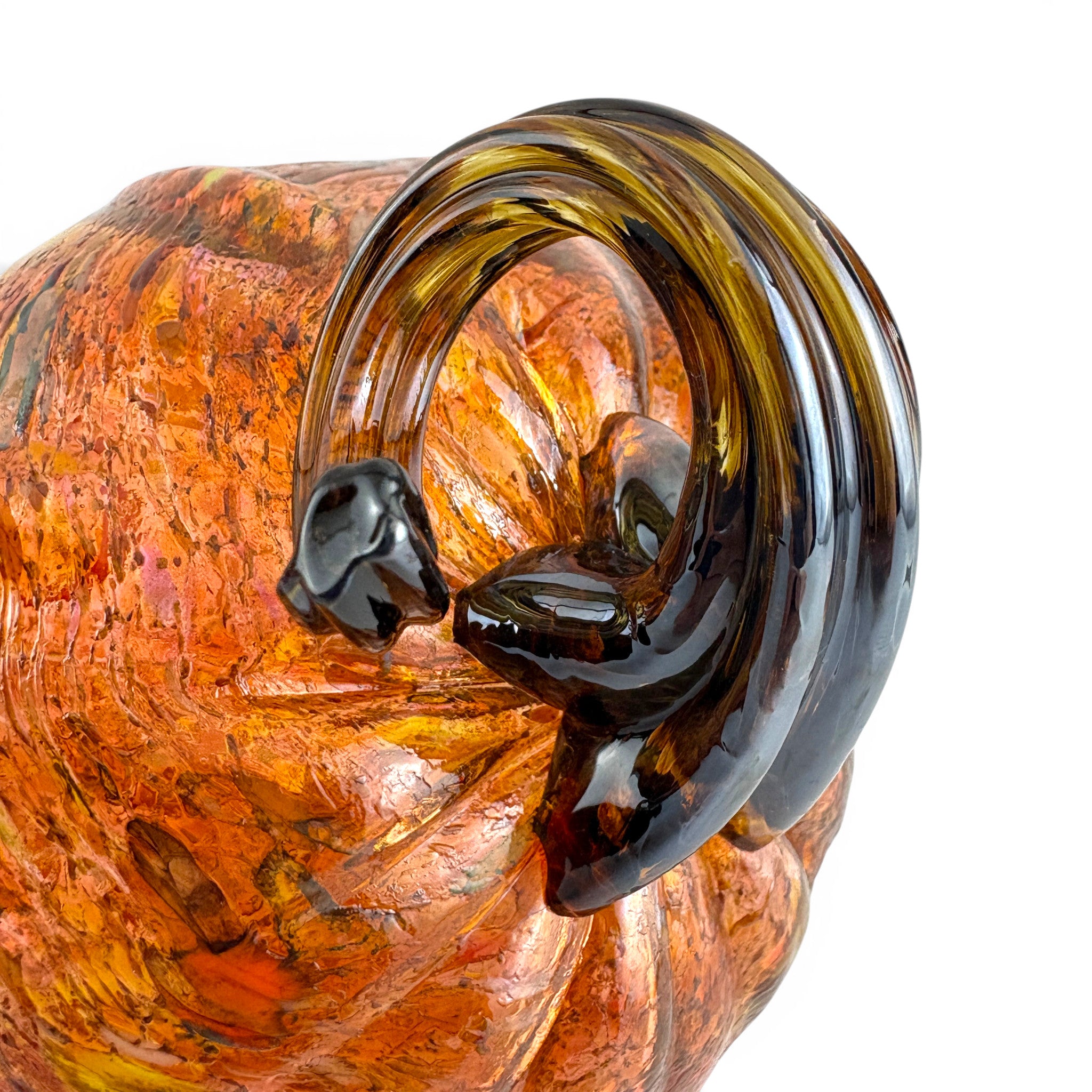A close-up of a decorative glass pumpkin featuring a glossy dark brown and amber swirl on the head and neck over a textured orange and yellow glass body, set against a plain white background.