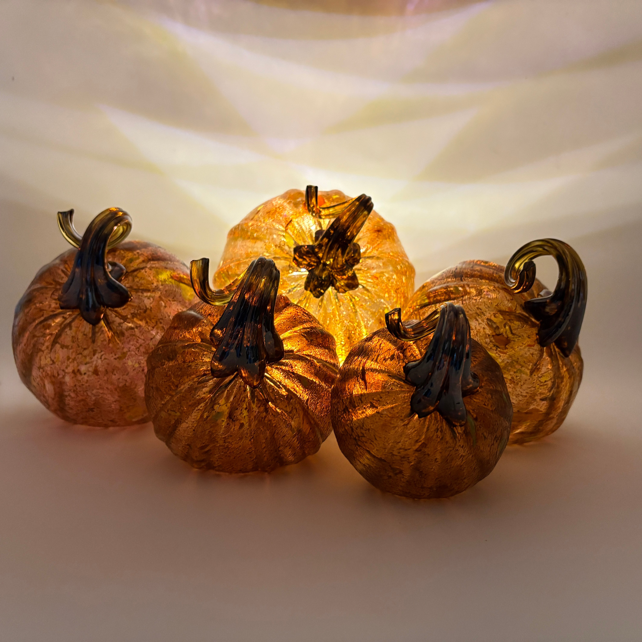 Five decorative glass pumpkins arranged together, glowing with warm amber light from within. The pumpkins have textured, metallic-looking orange and brown glass bodies with dark brown to black stems, set against a softly lit cream background.
