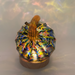 A decorative glass pumpkin with an iridescent, rainbow-reflective surface and a ribbed amber-brown stem, displayed on an LED light with a round wooden base. 