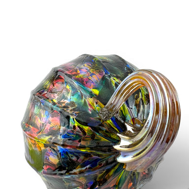 A close-up of a faceted glass container with a curved handle, featuring an iridescent finish that reflects multiple colors including green, blue, yellow, pink, and purple. 