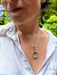 Person wearing a silver necklace with a pendant in front of green foliage