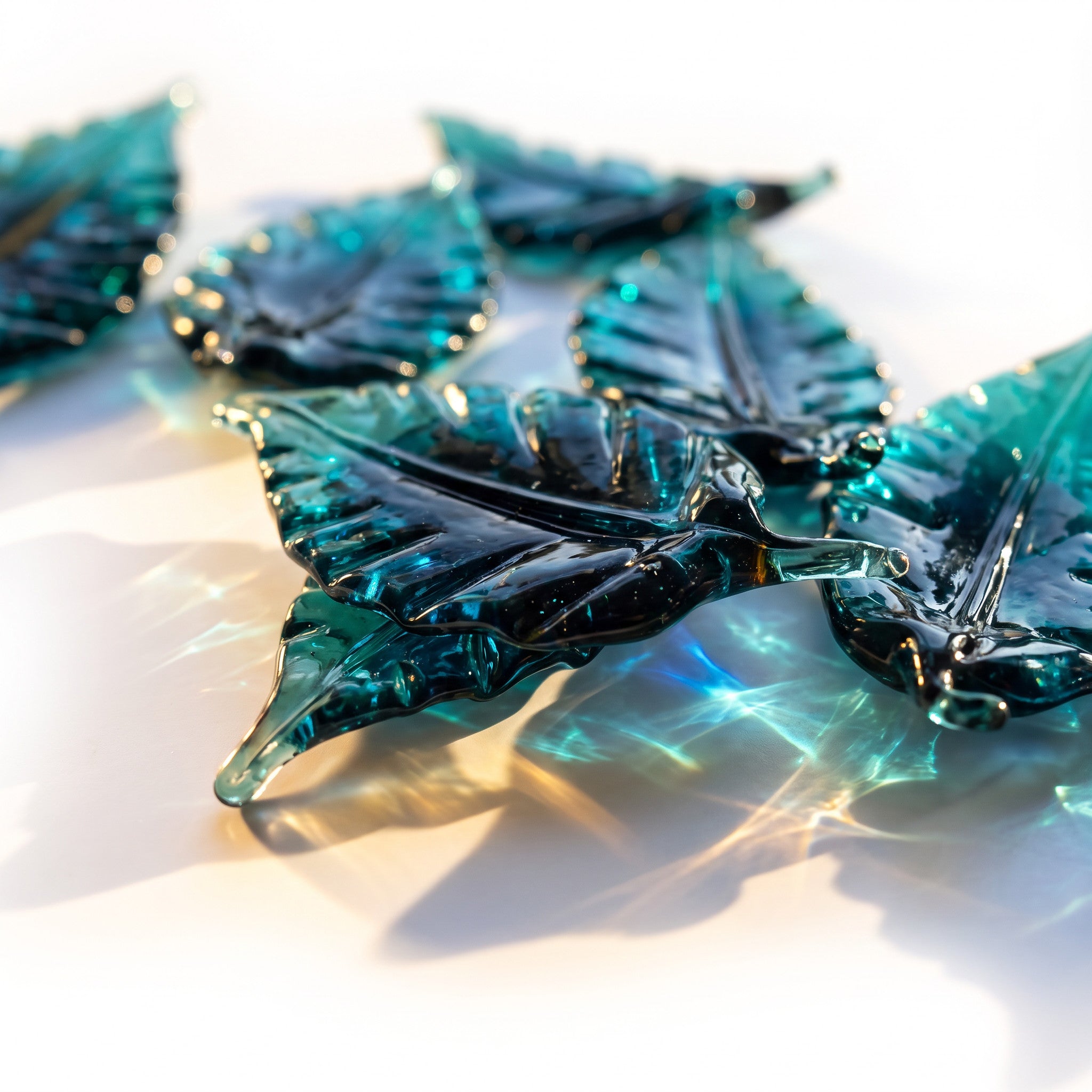 A close-up of translucent leaf-shaped glass sculptures in deep teal tones,   arranged on a bright white surface with sunlight filtering through.