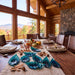 A dining table set in a rustic log cabin dining room, decorated with a beige woven runner, autumn leaves, acorns, small pumpkins, and several translucent teal glass leaf sculptures. Large windows offer a view of mountains, and a stone fireplace is visible in the background.