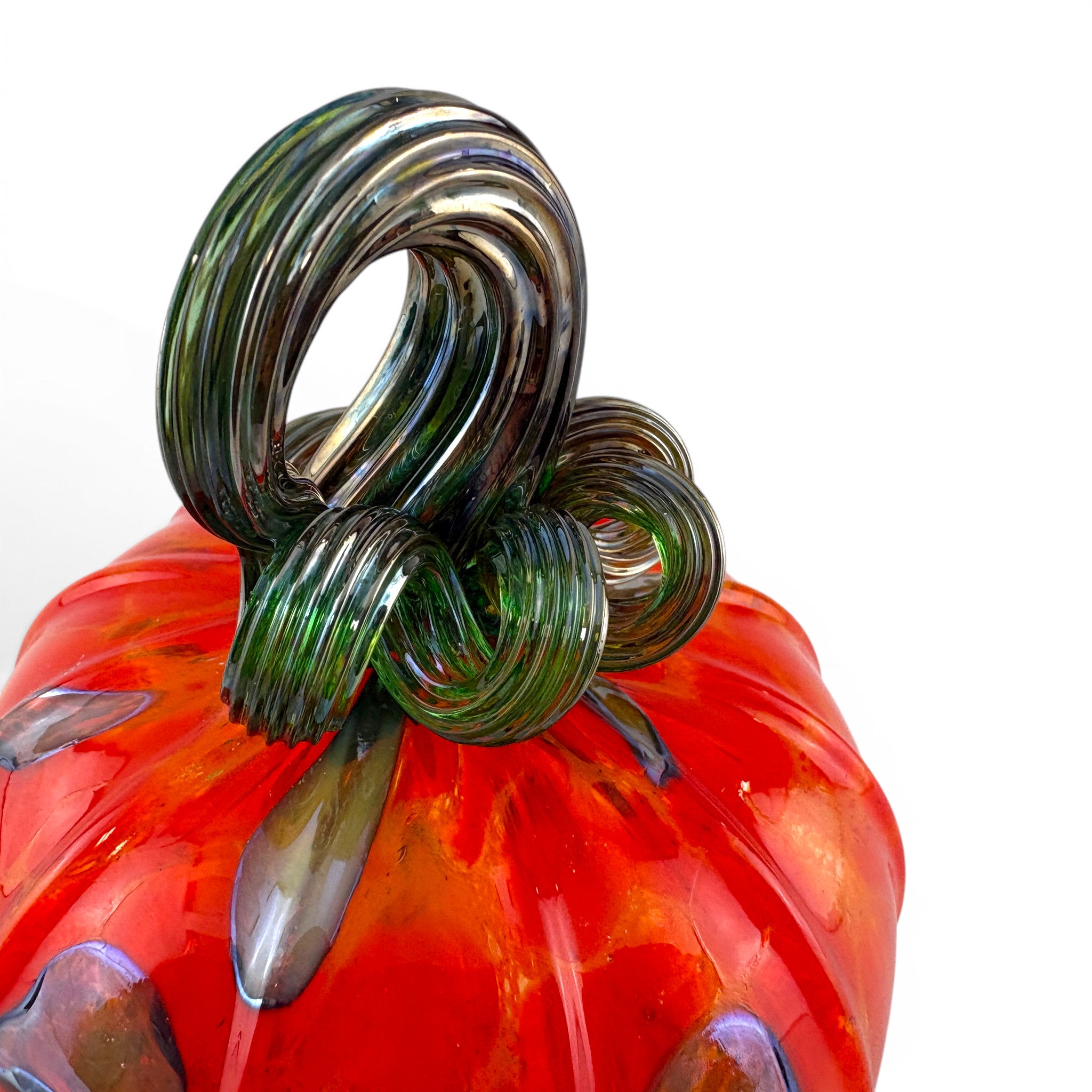 A close-up of a hand-blown glass pumpkin with a vibrant orange-red body, dark green stem, and subtle purple accents. 