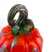 A close-up of a hand-blown glass pumpkin with a vibrant orange-red body, dark green stem, and subtle purple accents. 