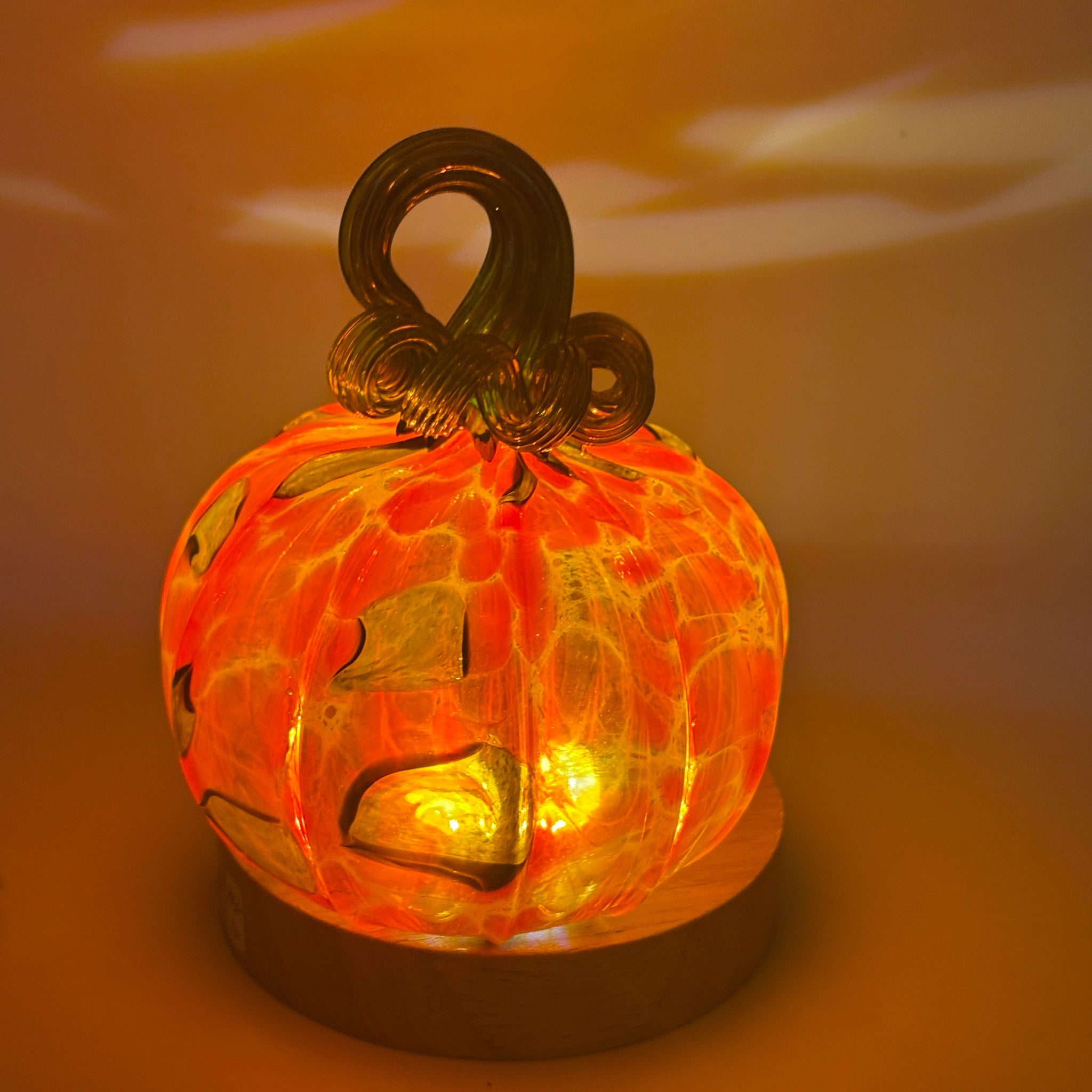 A handcrafted glass pumpkin glowing with warm orange and yellow light, featuring a dark glass stem, displayed on a round LED light with a wooden base. 
