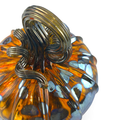 A close-up of a handcrafted glass pumpkin with a ribbed amber-orange body, dark gray smoky glass detailing, and a matching dark gray stem. 