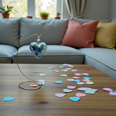 Decorative heart-shaped hand-crafted glass ornament on a stand with colorful heart-shaped confetti on a wooden table, in front of a gray sofa with cushions.