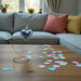 Decorative heart-shaped hand-crafted glass ornament on a stand with colorful heart-shaped confetti on a wooden table, in front of a gray sofa with cushions.