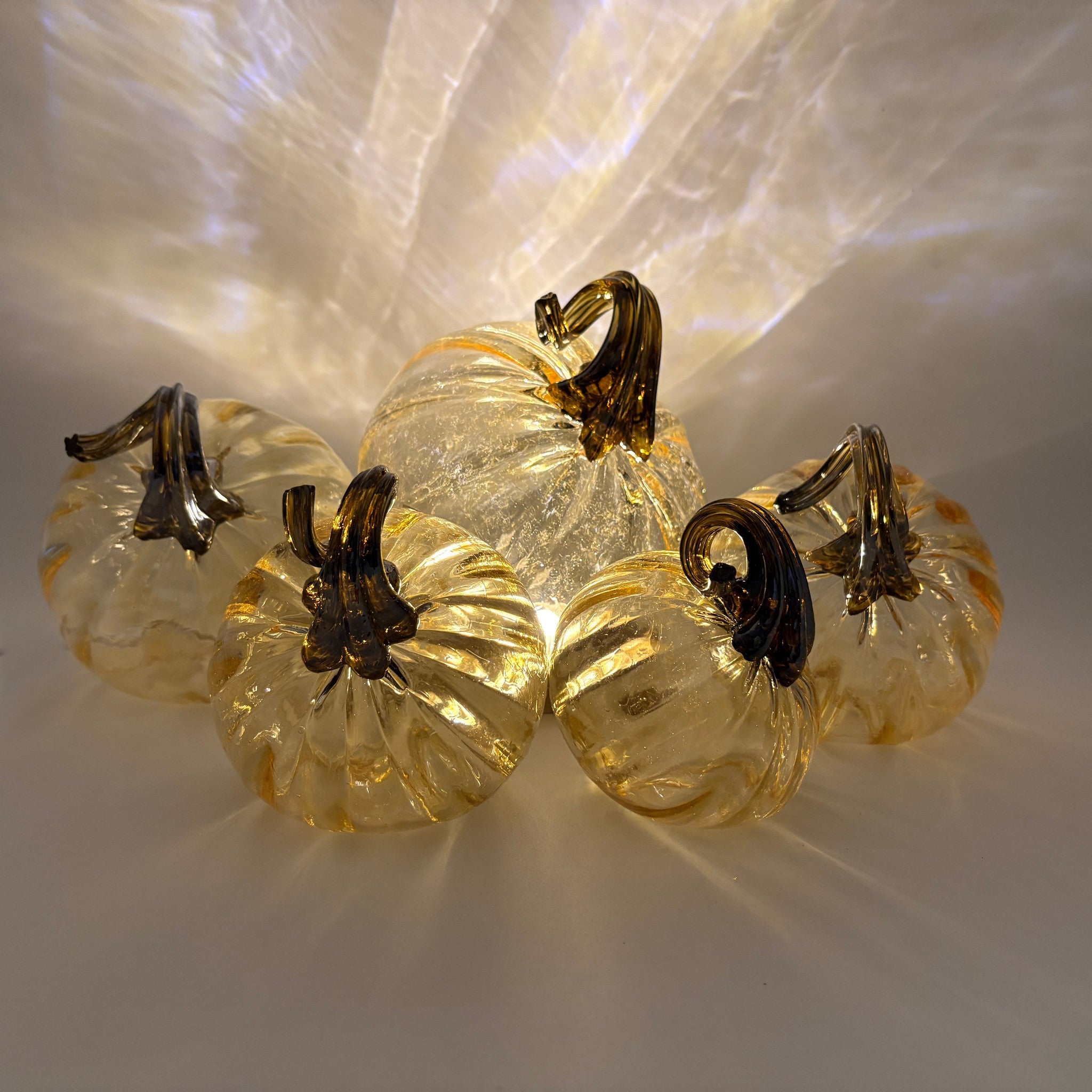 Five small clear glass pumpkins with amber-yellow tint and dark brown stems, arranged together on a light surface with warm light reflections.