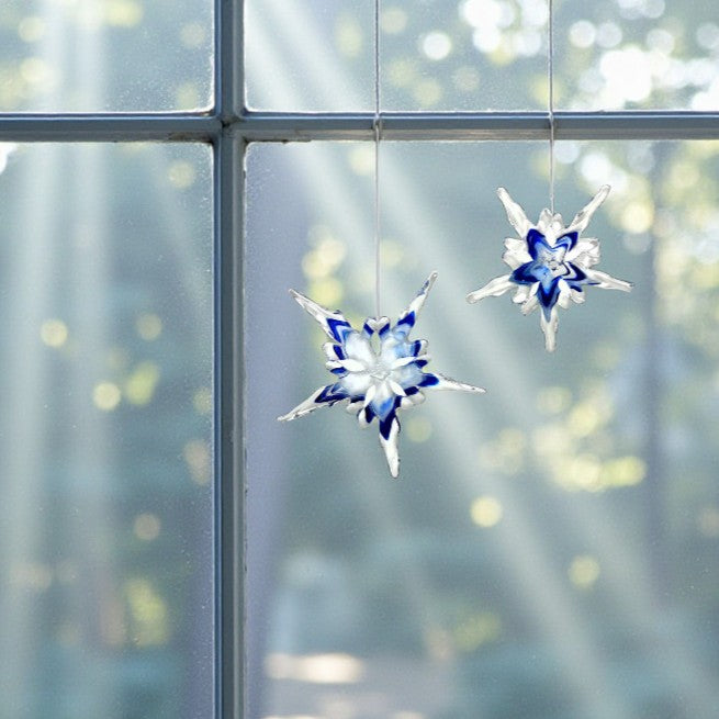 Two decorative handcrafted glass snowflakes hanging in front of a window with a blurred greenery background.
